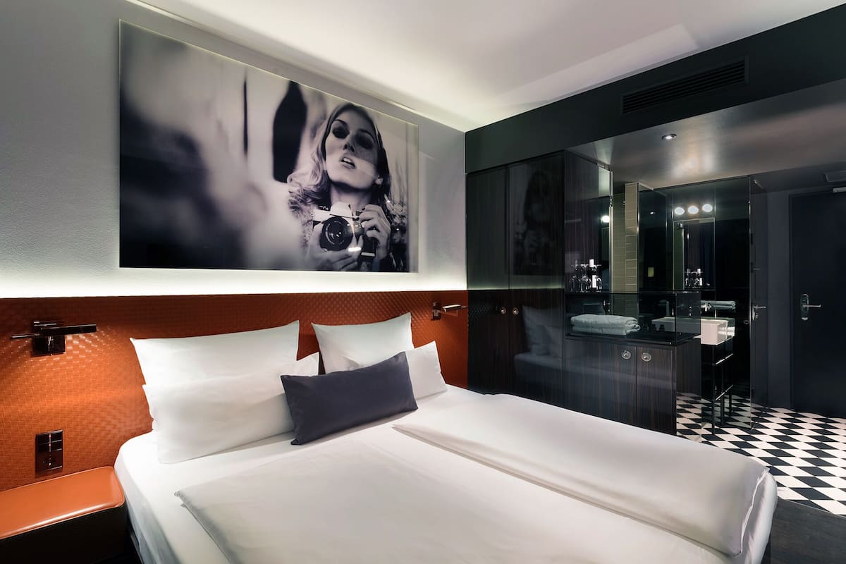 A modern hotel room with a large bed, a bathroom with a mirror, and a checkered floor.
