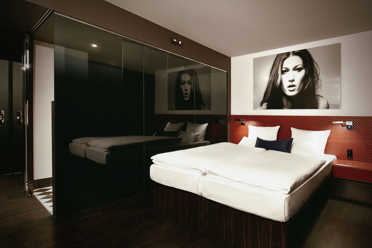 A modern bedroom with a large bed, a glass wardrobe, and a red accent wall.