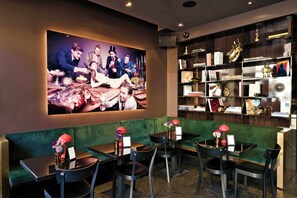 Un restaurante con asientos tapizados en verde, mesas y sillas negras. Se ve un televisor de gran tamaño montado en la pared mostrando una foto grupal.
