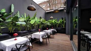 Un área de comedor al aire libre con mesas y sillas, rodeada de plantas grandes y lámparas colgantes.