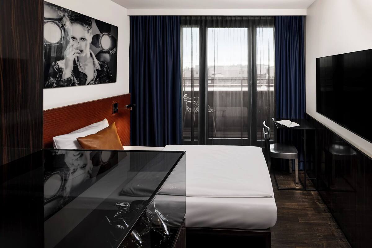 A modern hotel room with a bed, a desk, a TV, and a balcony with curtains.