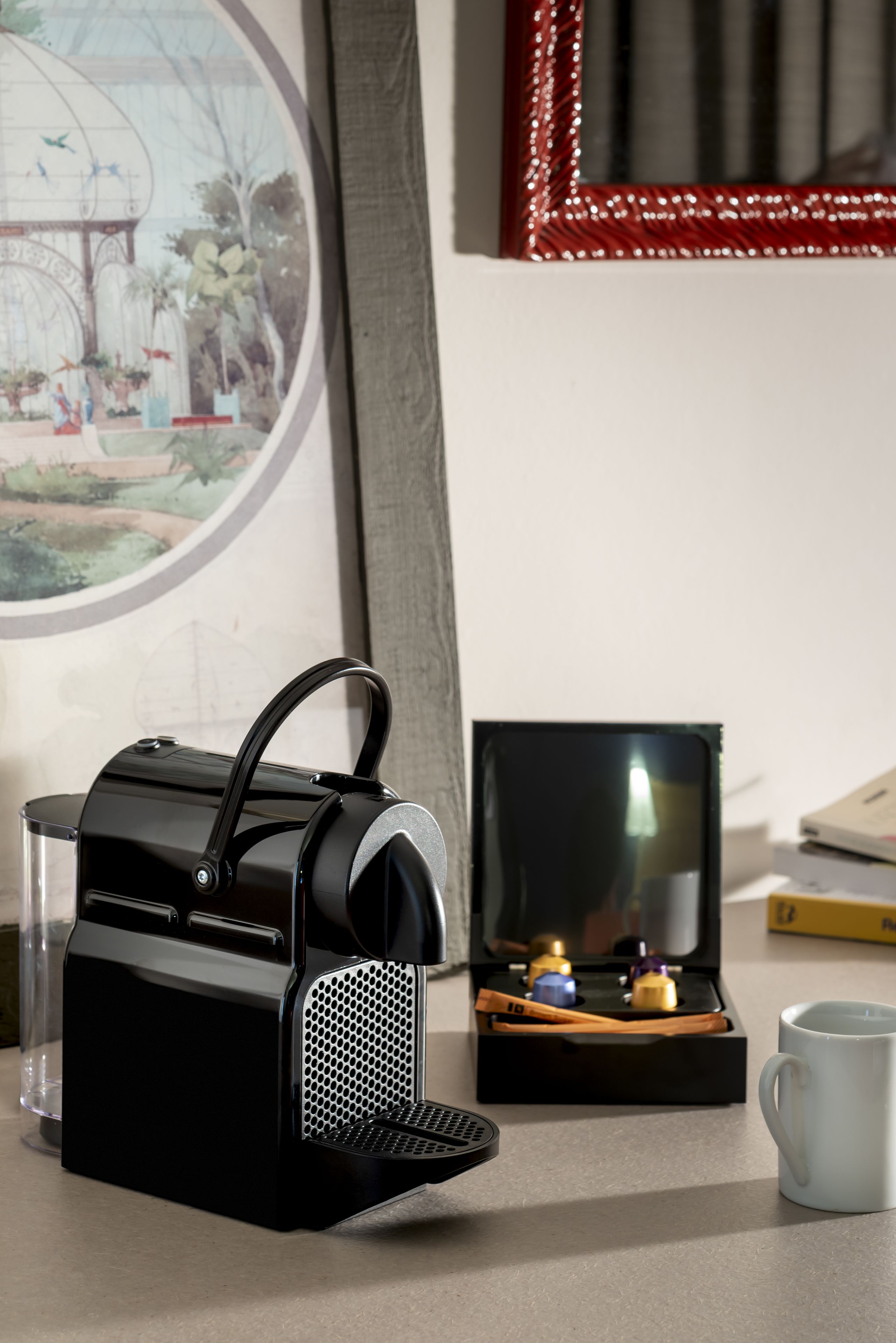 A black portable speaker on a table, a clear glass, a white mug, a framed picture, and a small box with items.