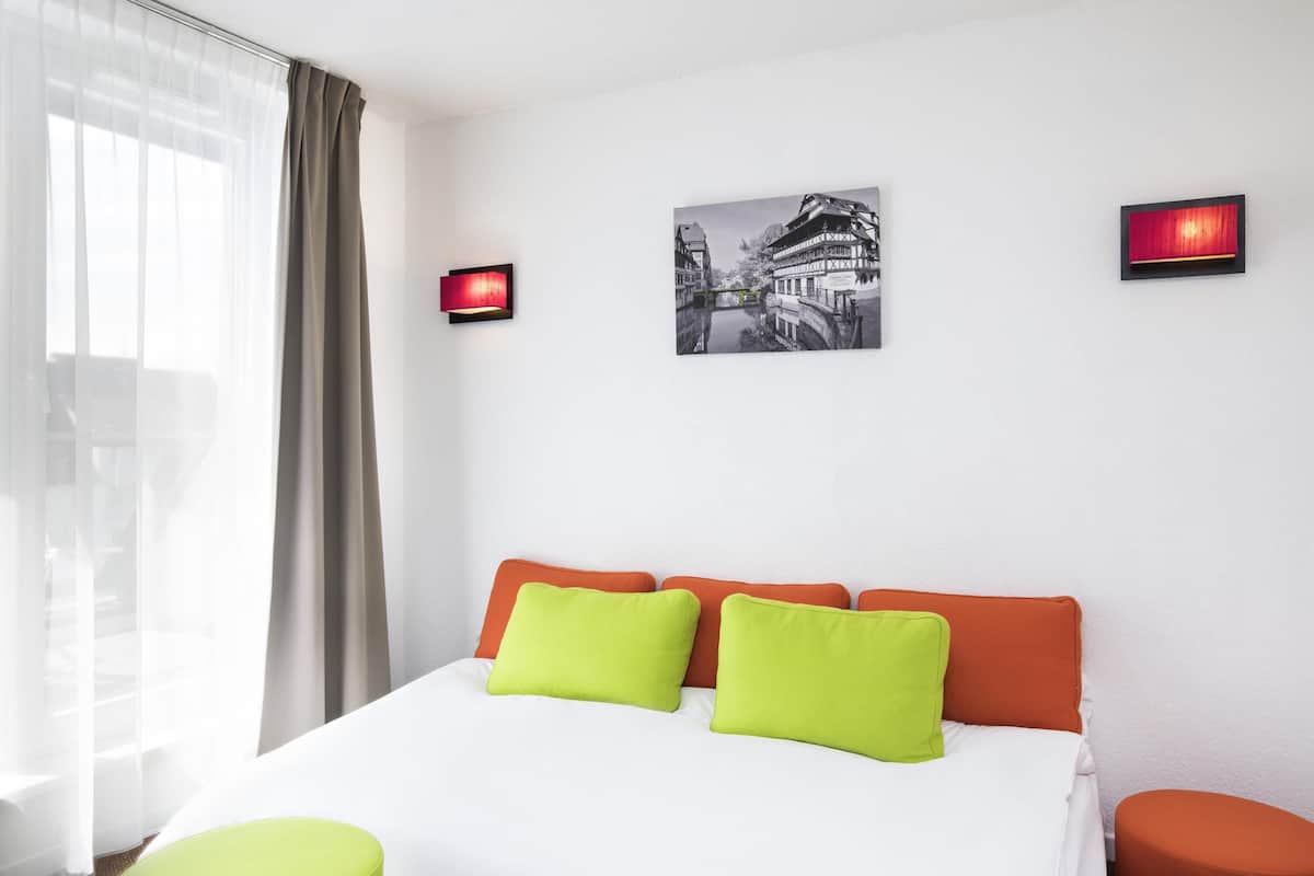 Ein Hotelzimmer mit Bett, zwei Kissen, einem Fenster mit Vorhängen, einem Wandbild und zwei Wandleuchten.