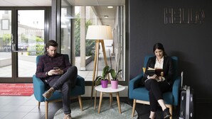 Two people sitting in a modern lobby with a 'HELLO' sign, a lamp, and a suitcase.