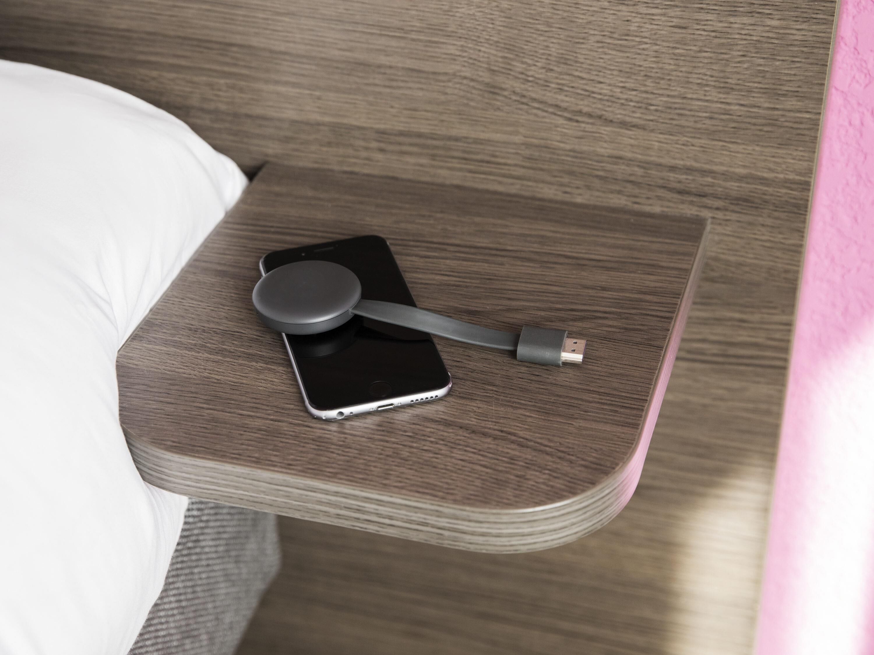 Un téléphone intelligent posé sur une table de chevet en bois, avec un câble USB et un appareil circulaire, probablement une station de chargement.