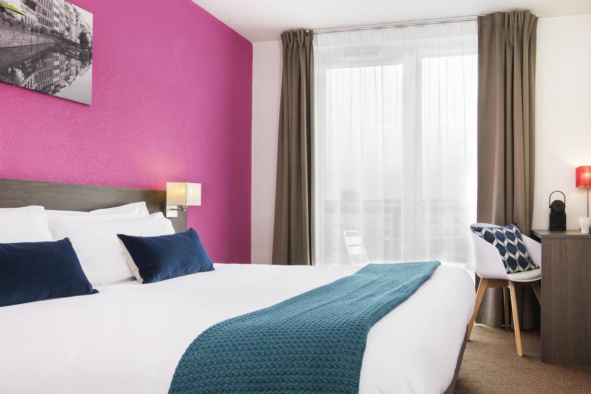 Ein Hotelzimmer mit einem großen Bett, einem Schreibtisch mit Stuhl, einem Fenster mit Vorhängen und einem Wandbild.