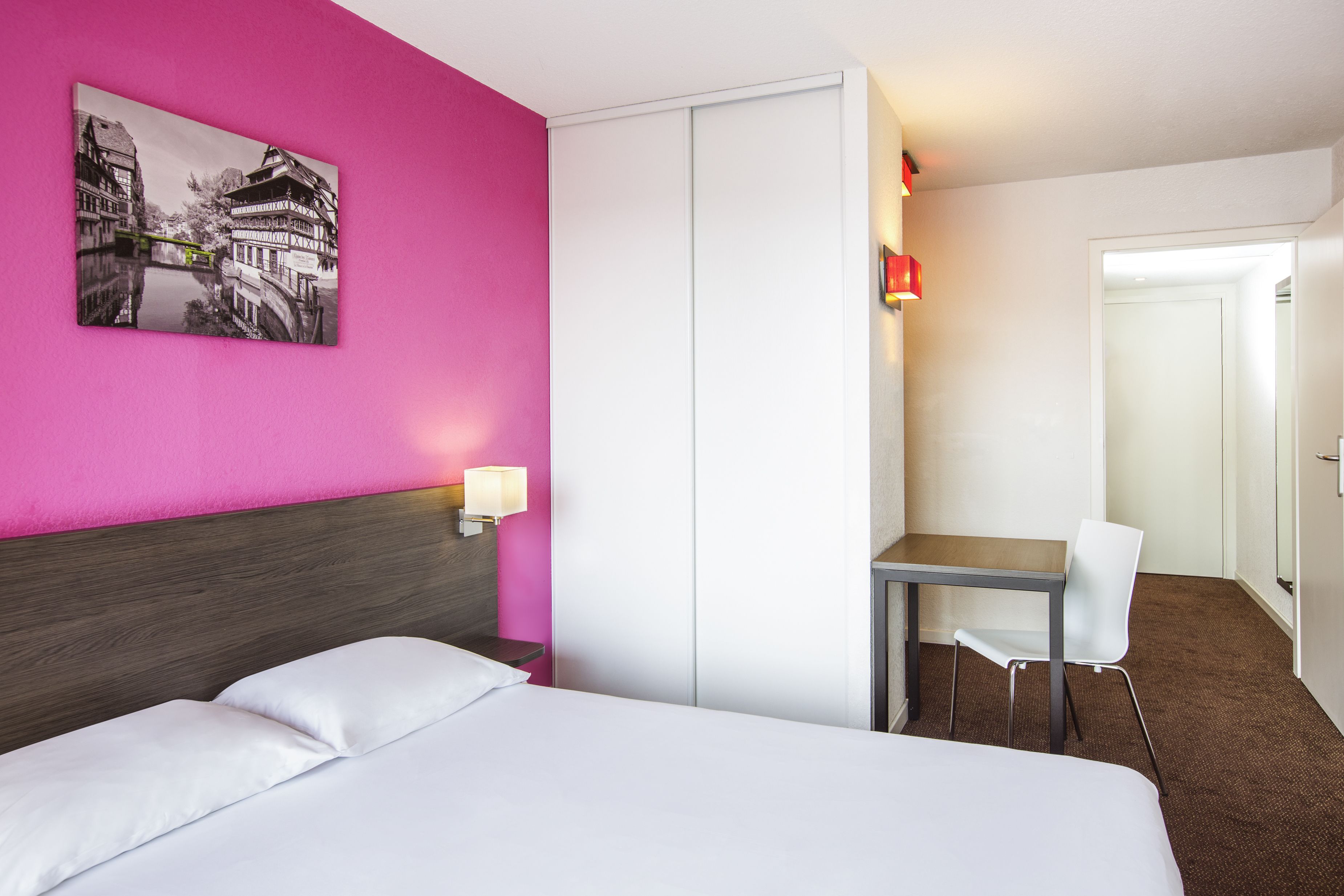 A hotel room with a bed, a desk, a chair, a nightstand, a lamp, and a wardrobe.