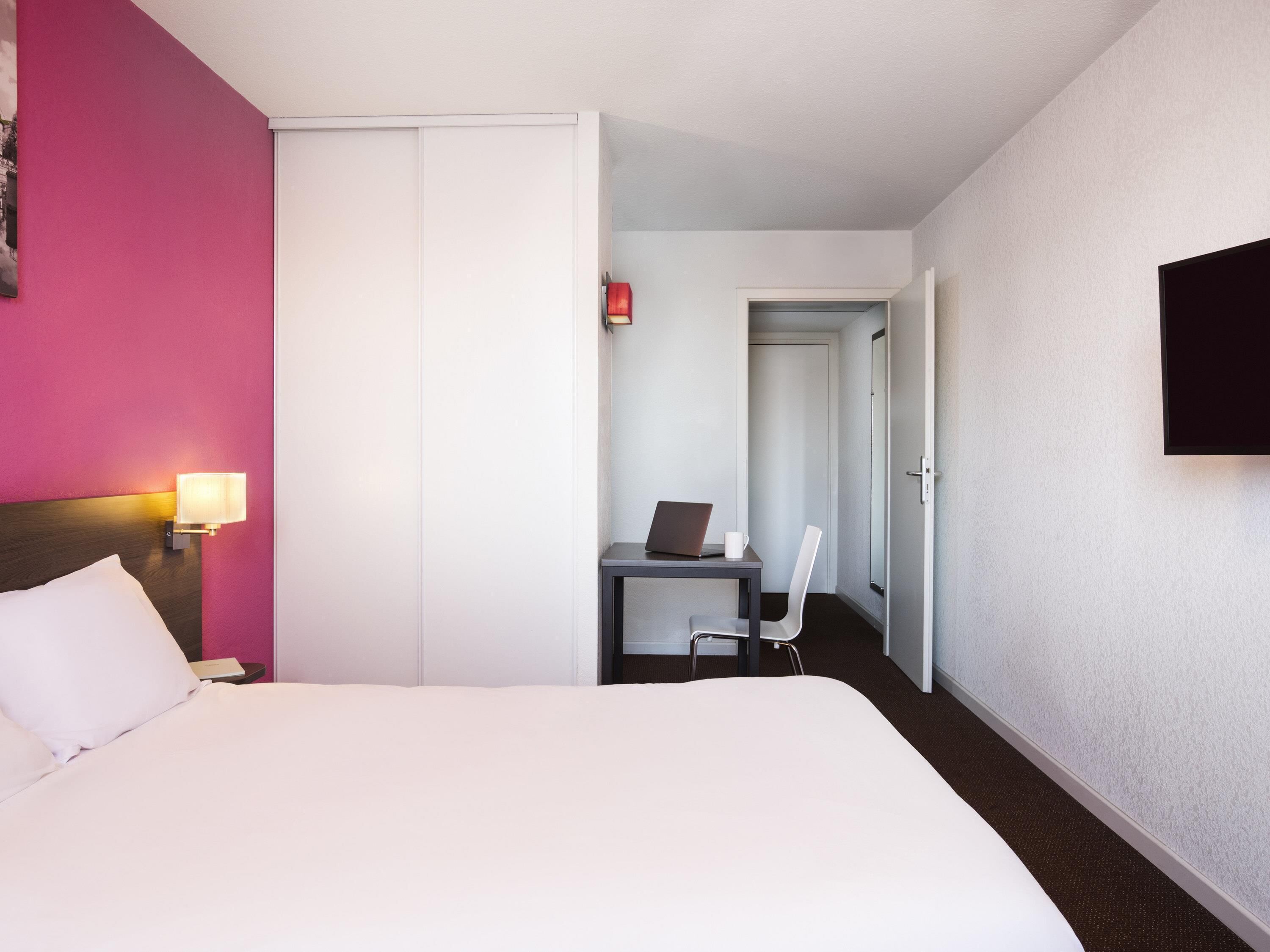 Una habitación de hotel con una cama, un escritorio con una computadora portátil, una silla y un televisor en la pared.