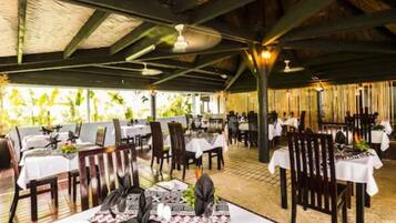 An outdoor restaurant with tables set for dining, featuring a thatched roof and natural light.