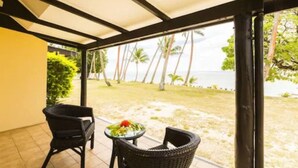 Un espacio de estar techado al aire libre con dos sillas de mimbre y una mesa pequeña, con vista a una playa con palmeras y el océano.