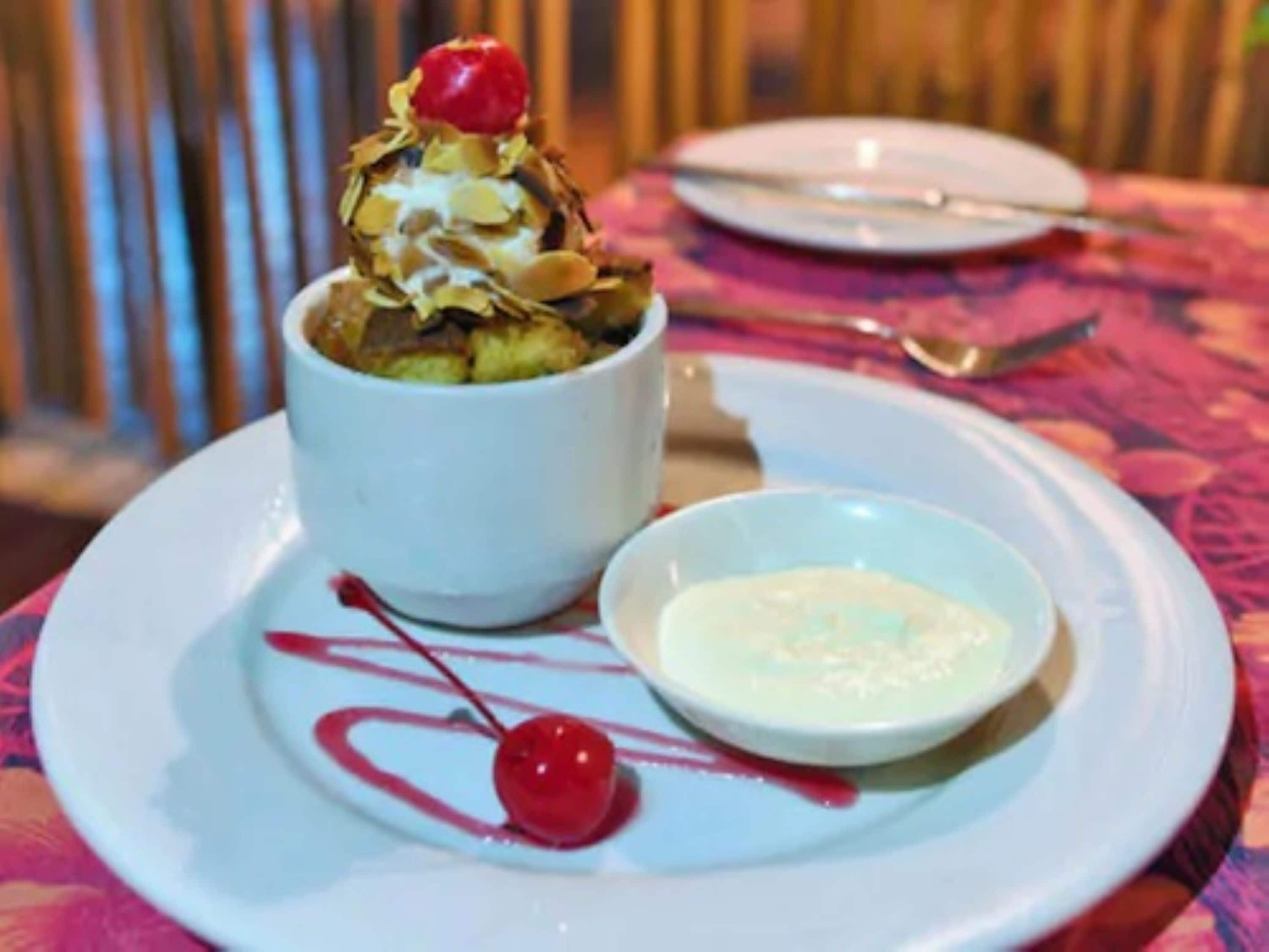 A dessert on a white plate with a cherry on top, a small bowl of sauce, and a spoon.