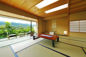 A tatami mat room with a low table, chairs, and a view of greenery and mountains.