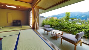 A tatami mat room with a view of mountains, a wooden table, two chairs, and a traditional Japanese cabinet.