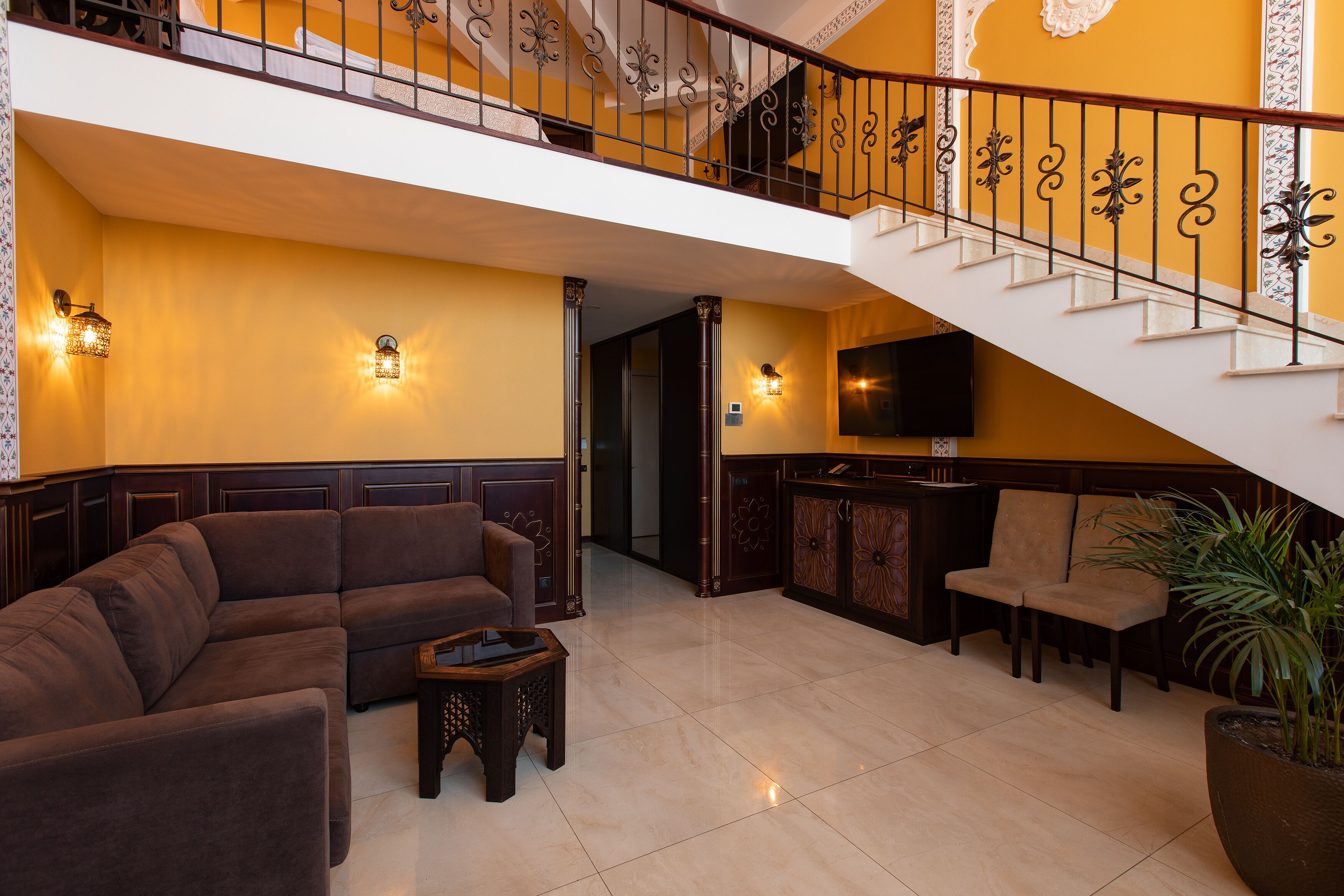 A spacious lobby with a large sectional sofa, a small coffee table, and a seating area with two chairs. A staircase with ornate railings leads to an upper level.