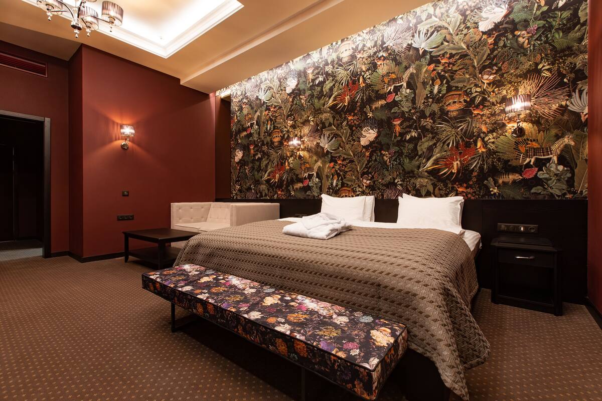 A hotel room with a large bed, a bench, a nightstand, and a wall with a floral mural.