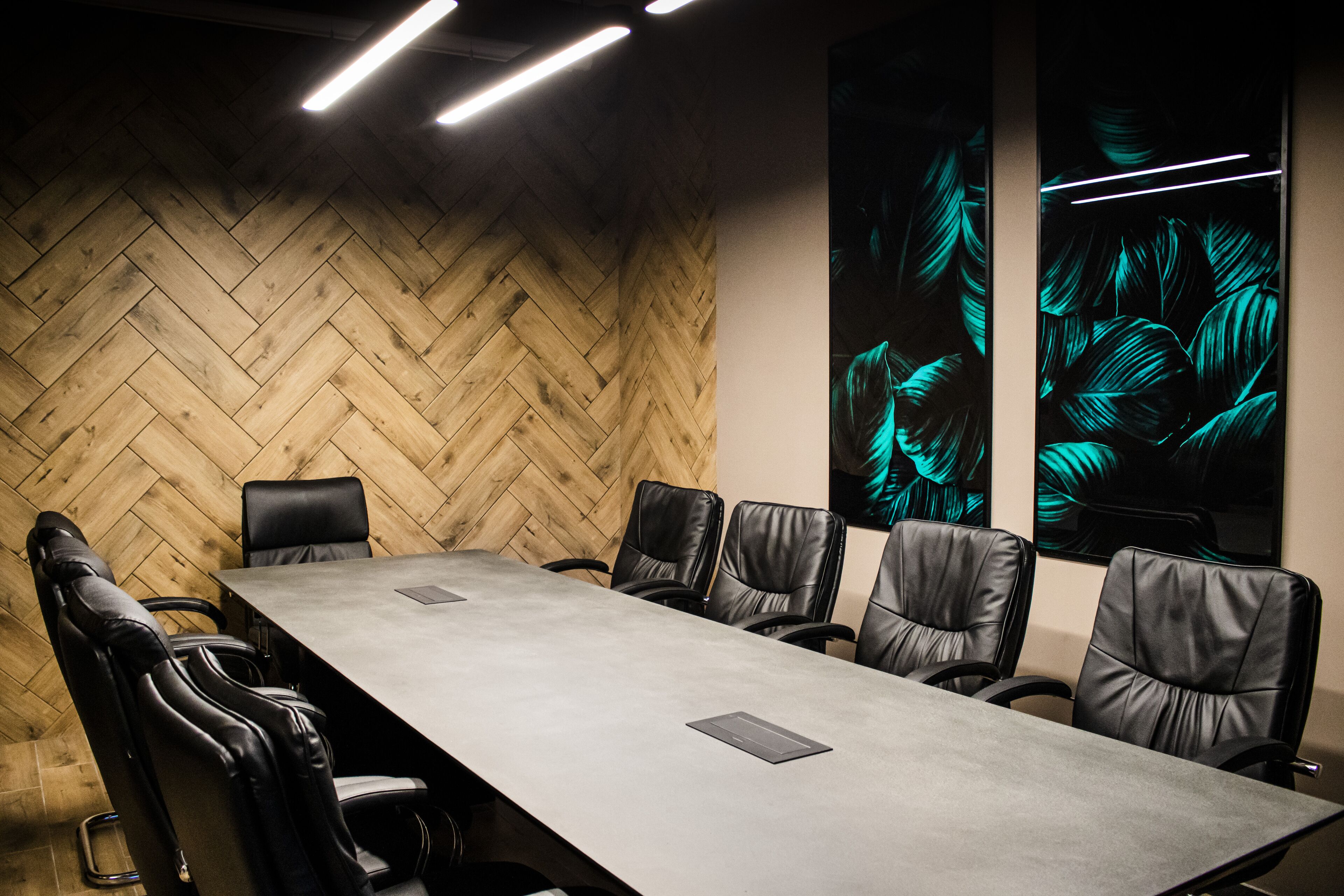 A conference room with a long table, six leather chairs, a herringbone wooden wall, and two illuminated wall art pieces.