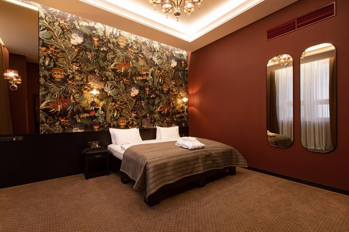 A hotel room with a large bed, a nightstand, a mirror, and a floral wallpaper.
