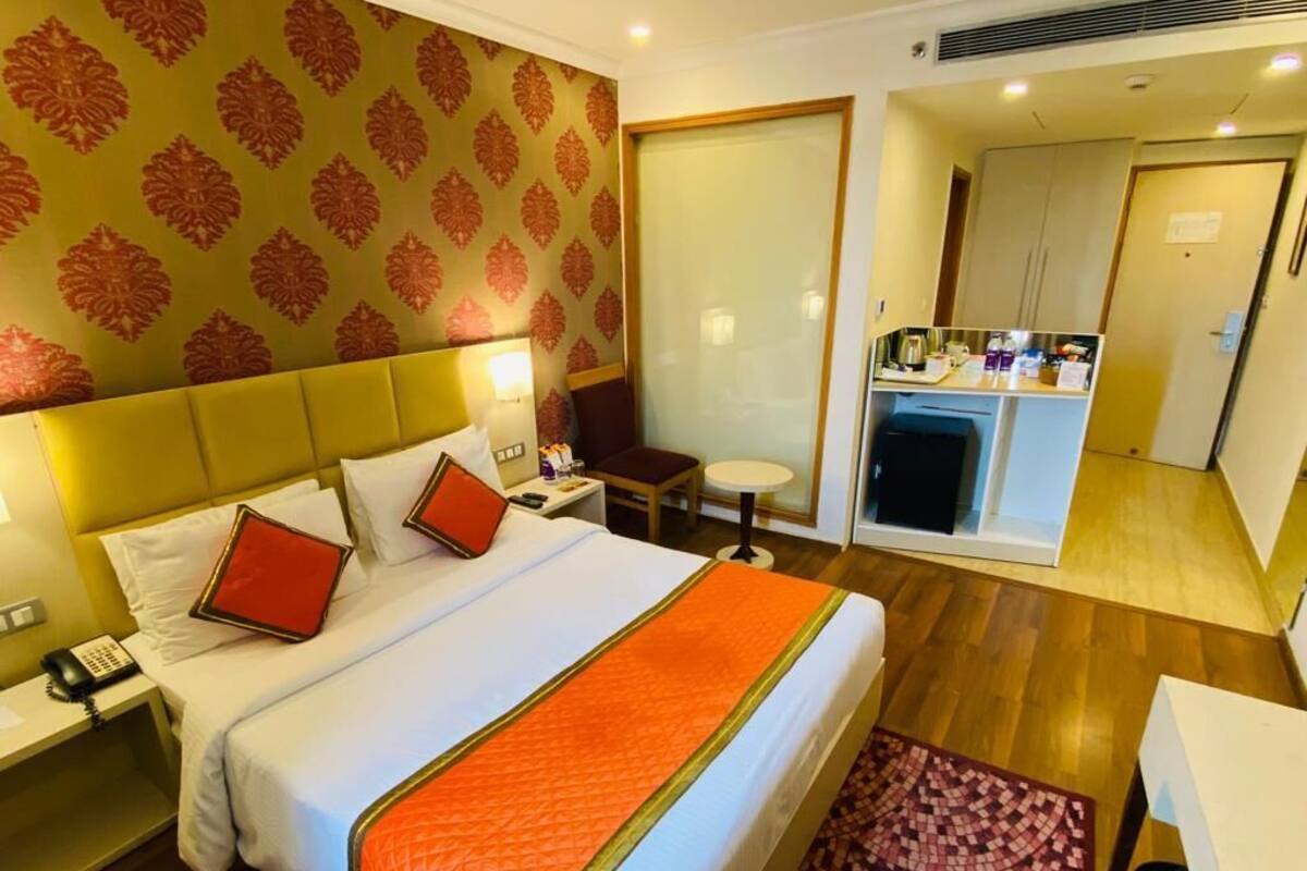 A hotel room with a large bed, a bedside table with a lamp, a small desk with a chair, and a minibar.