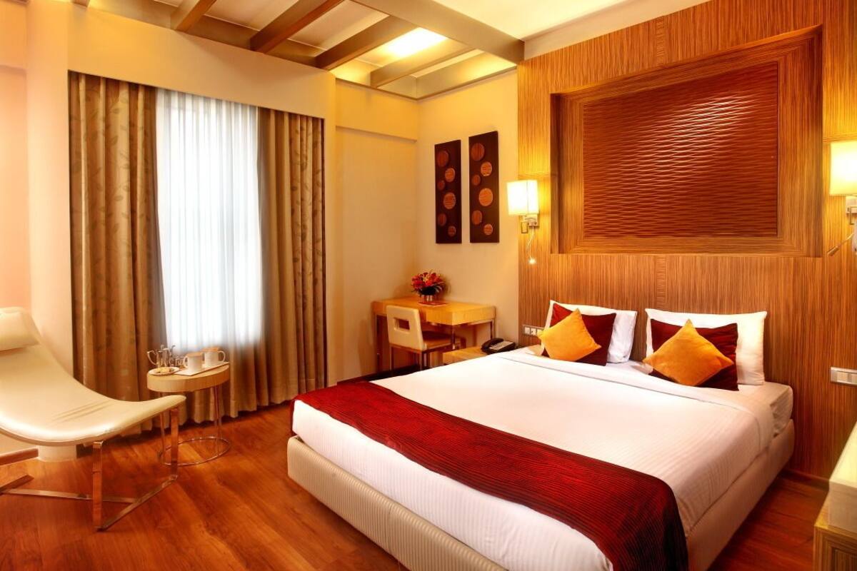 A hotel room with a large bed, a desk with a chair, a small table with a vase of flowers, and a wall-mounted lamp.