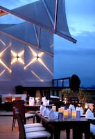 An outdoor dining area with tables set for two, candles, and a large canopy providing shade.