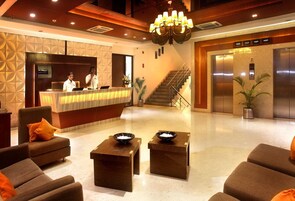 A hotel lobby with a reception desk, two receptionists, a staircase, and an elevator.
