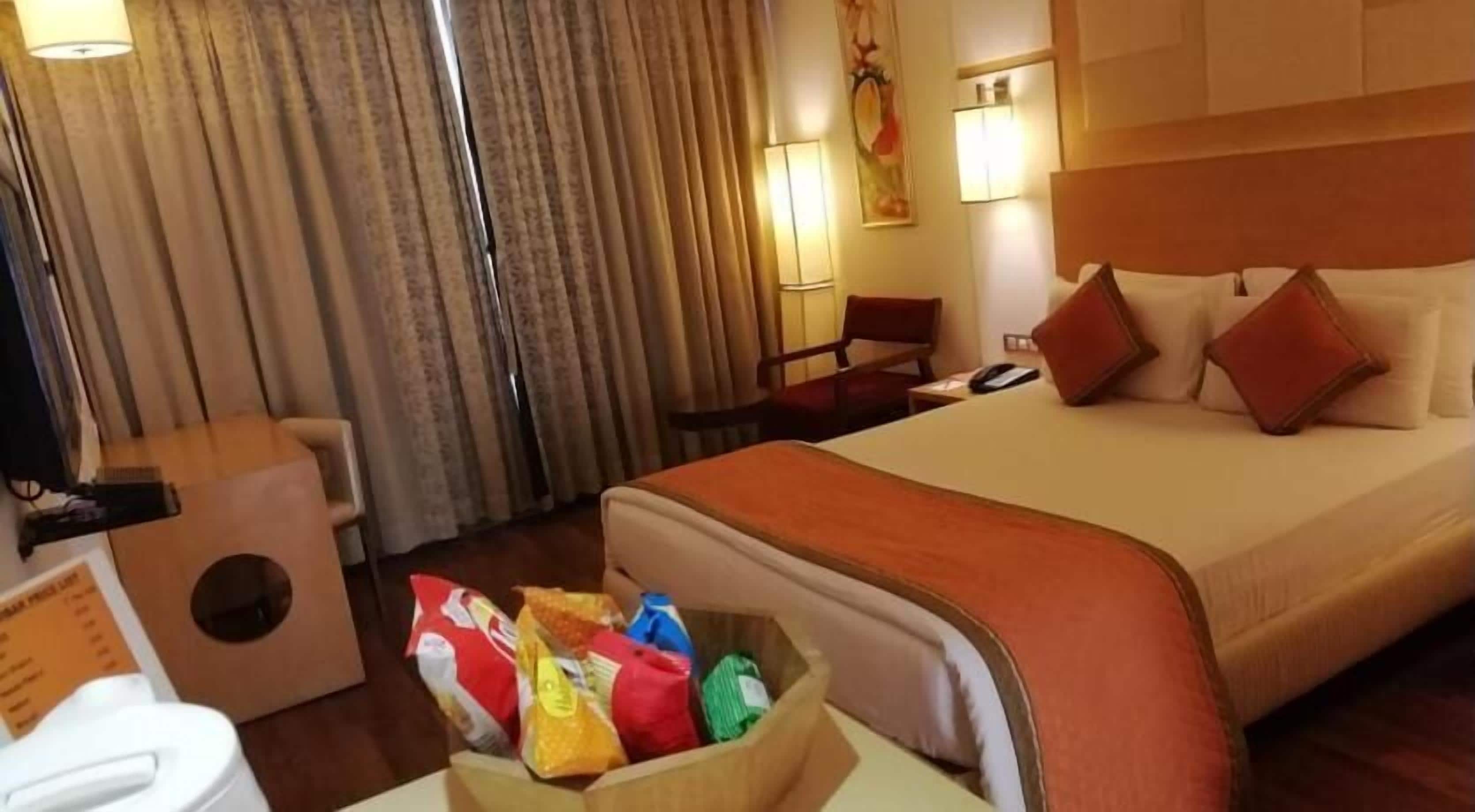 A hotel room with a large bed, a wooden headboard, a bedside table with a lamp, a chair, and a small table with snacks.