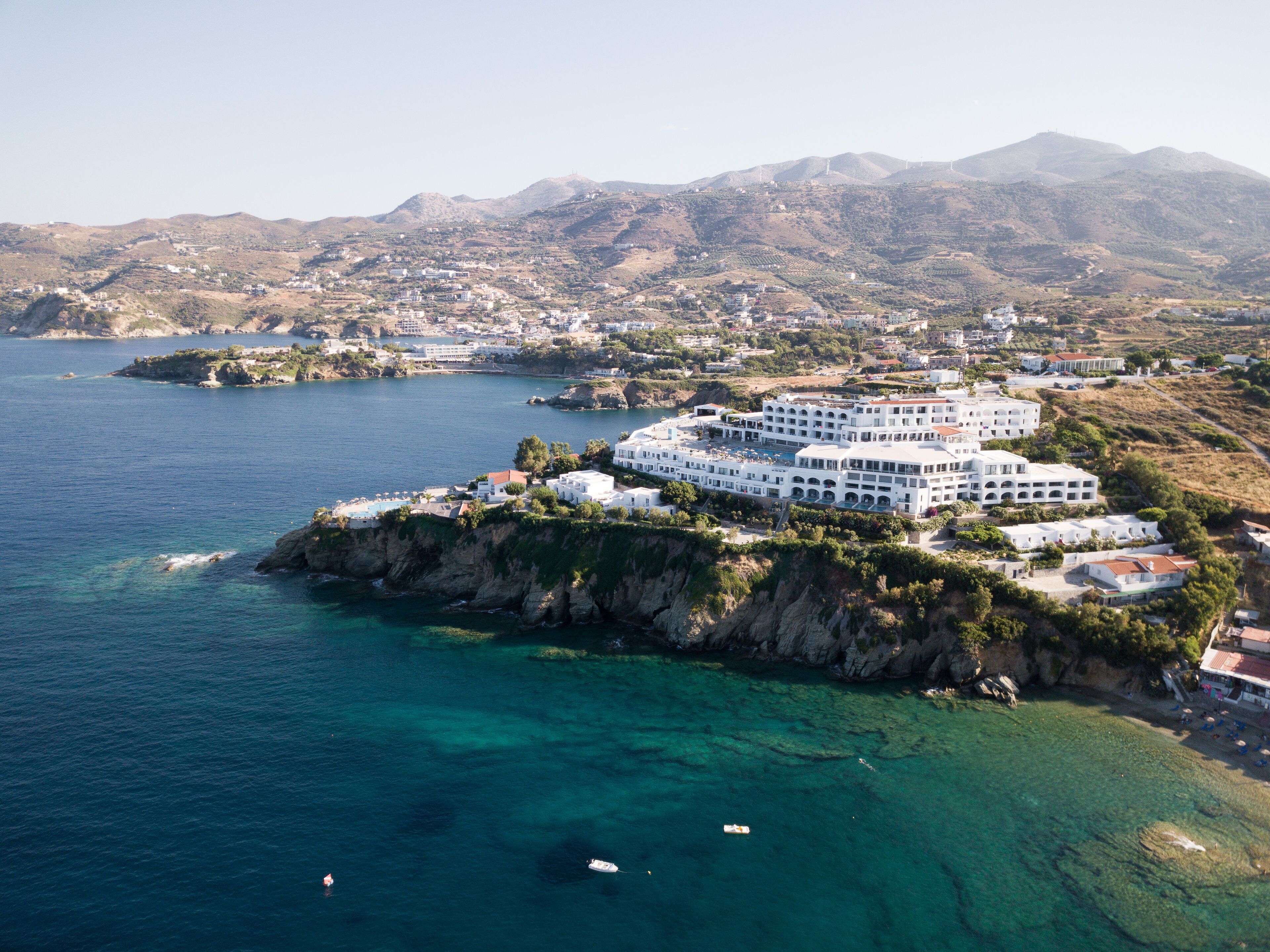 A coastal hotel with multiple white buildings, a rocky shoreline, and clear blue waters.