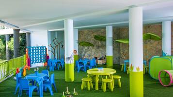 An indoor play area with green artificial grass, blue and yellow tables and chairs, and stone wall decorations.