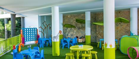 Un área de juegos interior con césped artificial verde, mesas y sillas azules y amarillas, y decoración de pared de piedra.