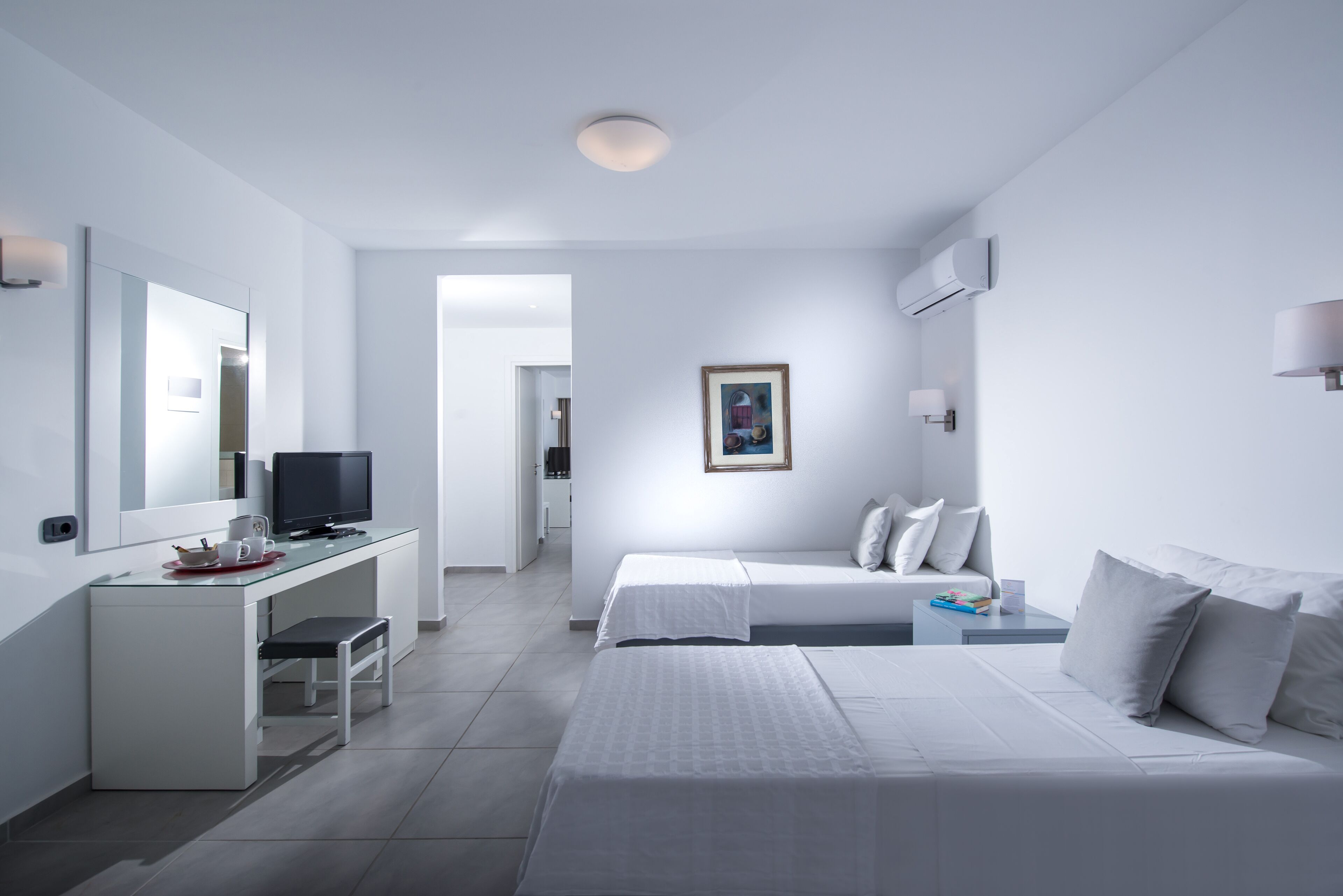 A modern hotel room with a white color scheme, a flat-screen TV, a desk with a chair, two beds, and a framed picture on the wall.