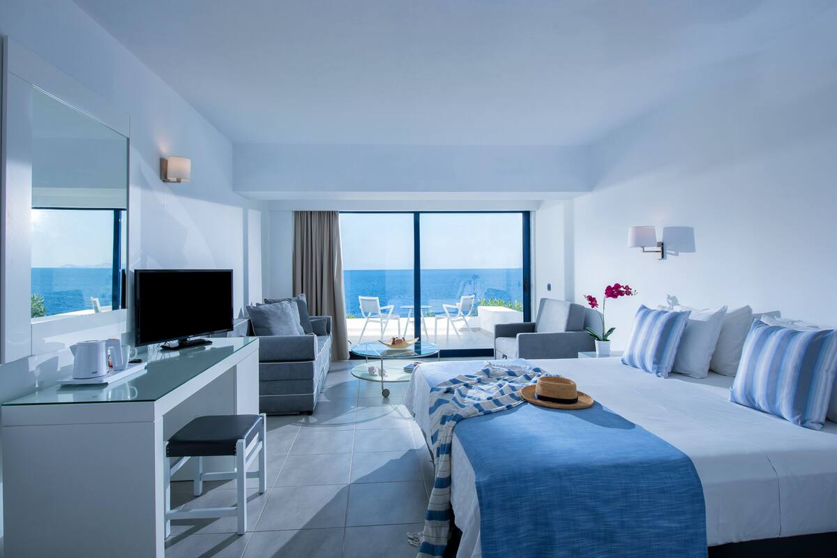 A modern hotel room with a large bed, a desk, a television, and a balcony with a view of the sea.