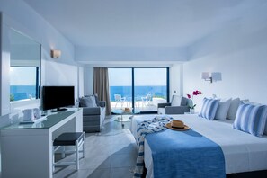 A modern hotel room with a large bed, a desk, a television, and a balcony with a view of the sea.
