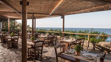 Un área de comedor al aire libre con suelo de piedra, mesas y sillas de madera, con vistas al mar.