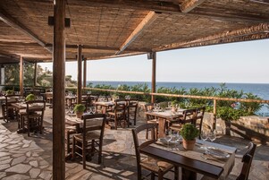 Un área de comedor al aire libre con suelo de piedra, mesas y sillas de madera, con vistas al mar.