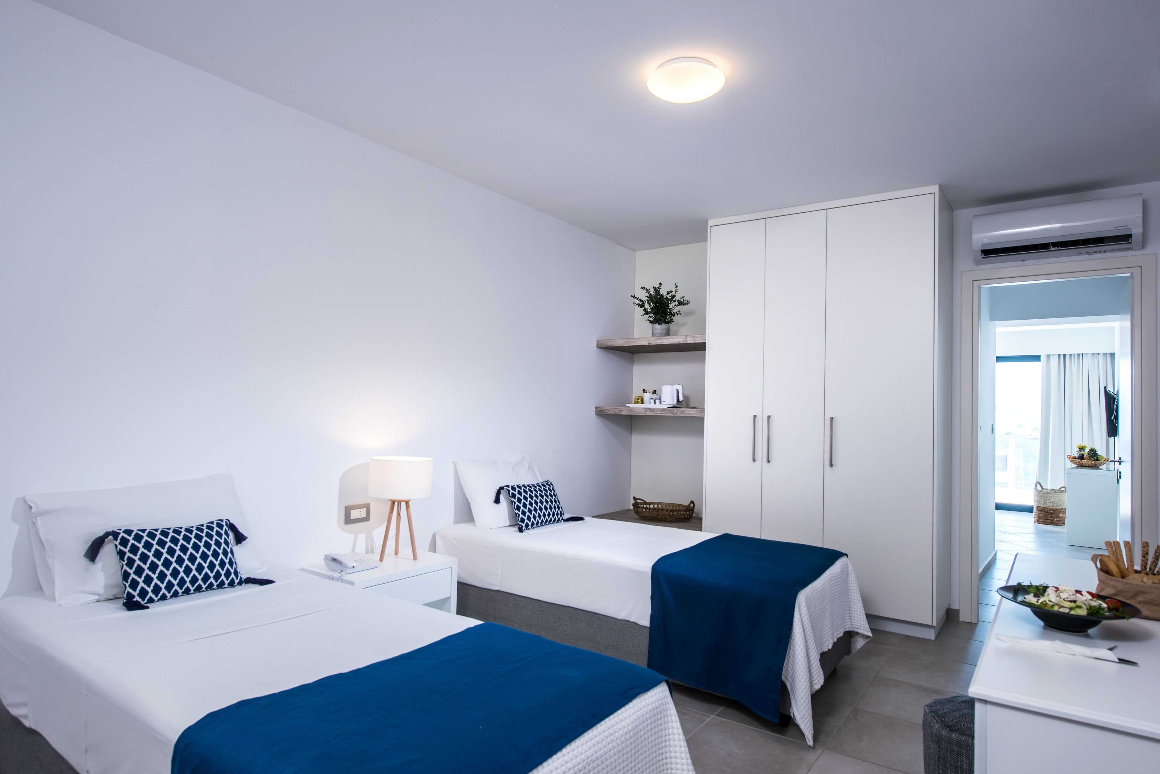 A hotel room with two beds, a desk, a chair, a small table with a bowl of fruit, a plant on a shelf, and an air conditioning unit.