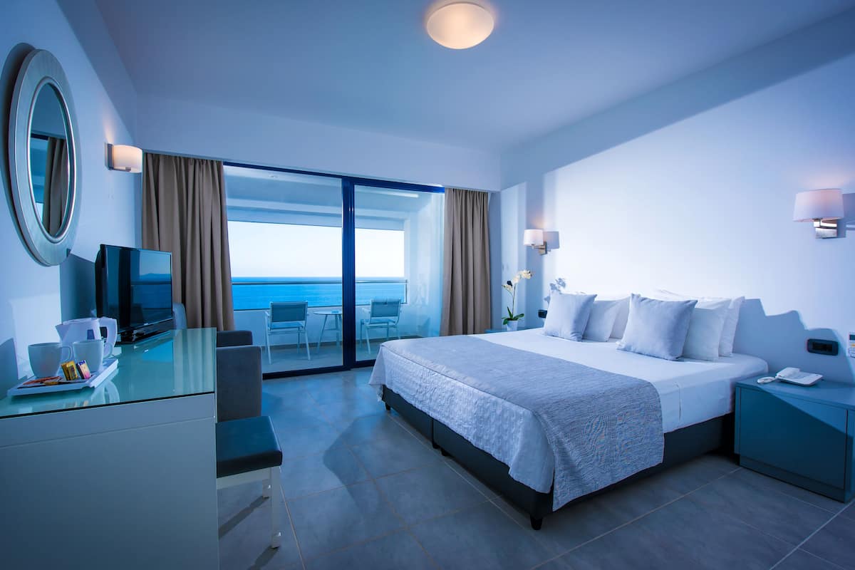 A modern hotel room with a large bed, a desk with a computer, and a view of the sea.