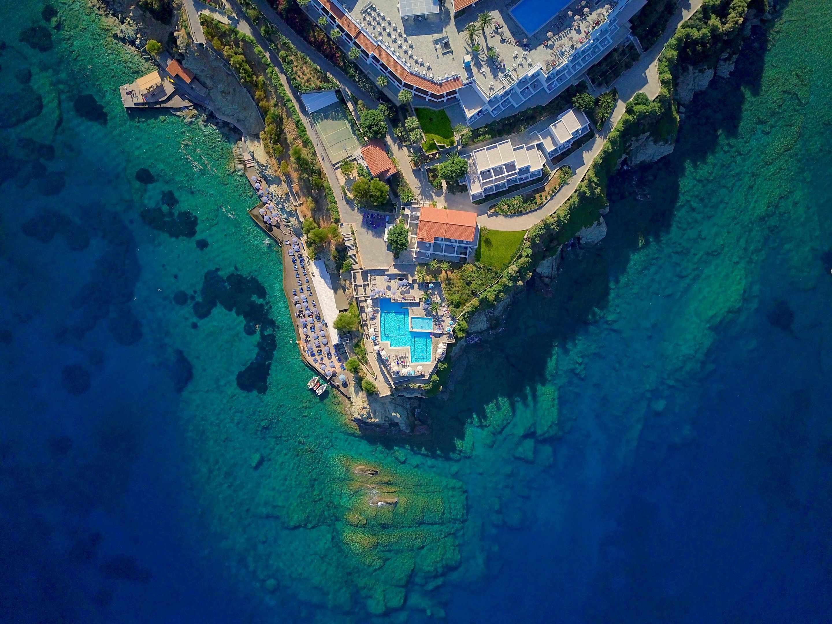 An aerial view of a coastal resort with a swimming pool, beachfront seating, and a cliffside location.