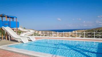 A swimming pool with two slides, a fenced area, and a view of the sea.