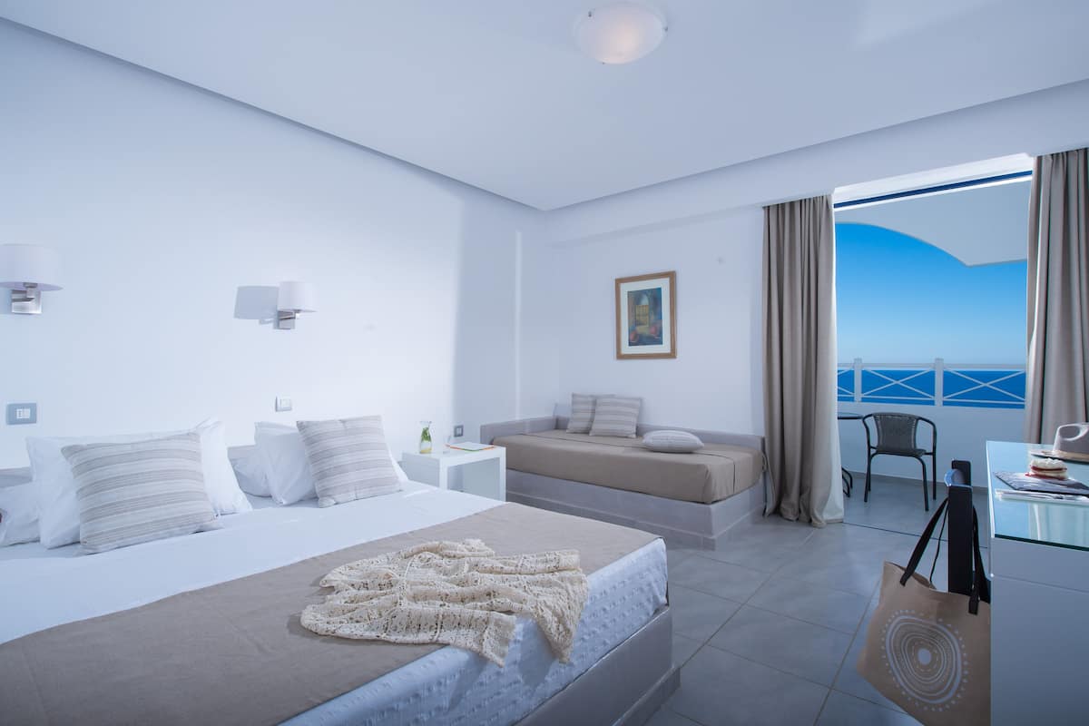 A modern hotel room with a large bed, a sofa bed, a balcony with a view, and a desk with a chair.