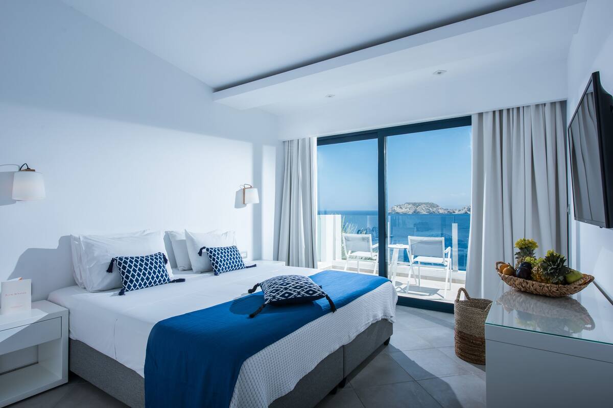 A modern hotel room with a large bed, a balcony with a view of the sea, and a wicker basket with fruits on the nightstand.