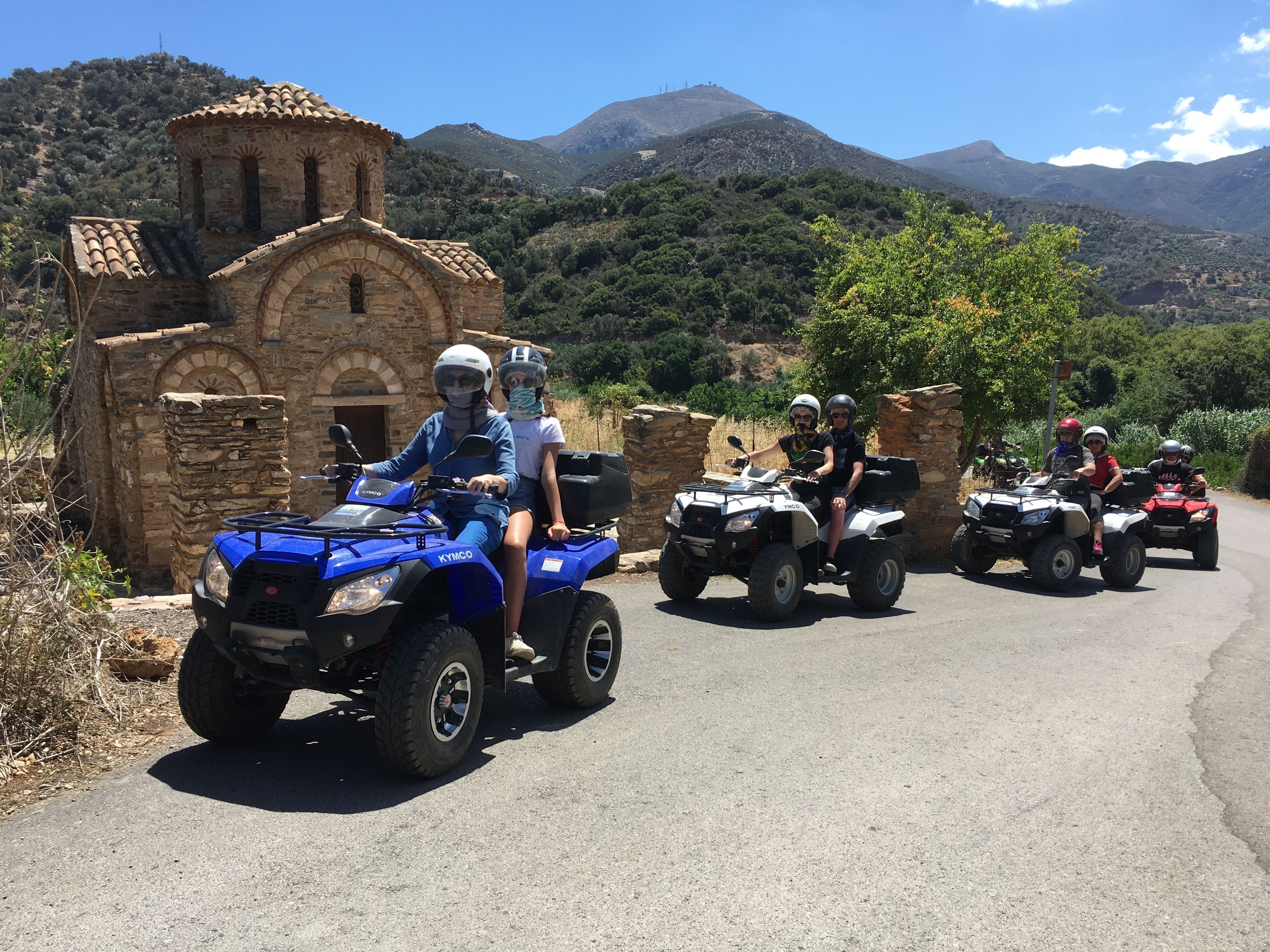 A group of people on ATVs on a road with a stone building and mountains in the background.