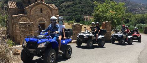 A group of people on ATVs on a road with a stone building and mountains in the background.