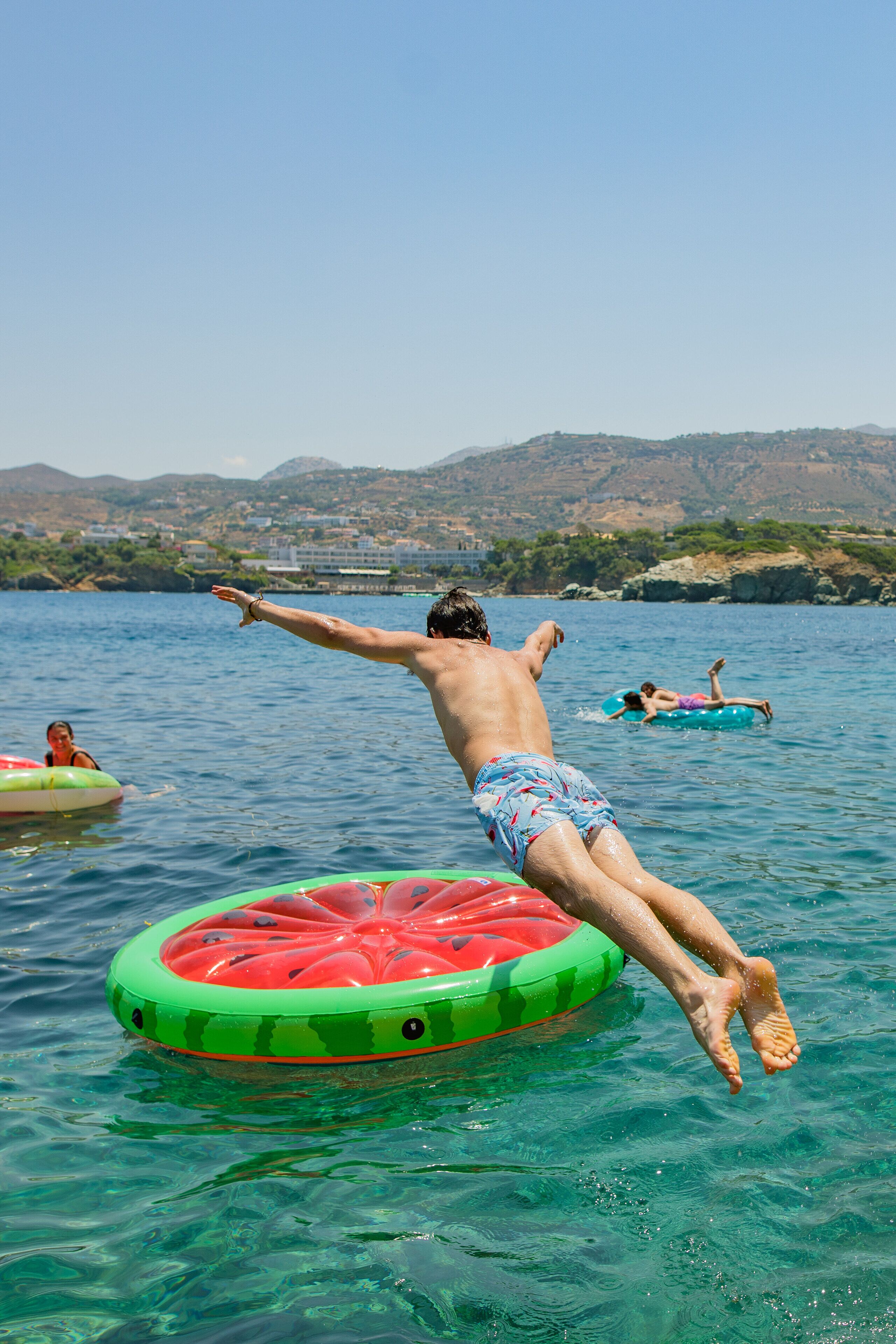A man jumping off a watermelon float into a lake with clear water and distant hills.