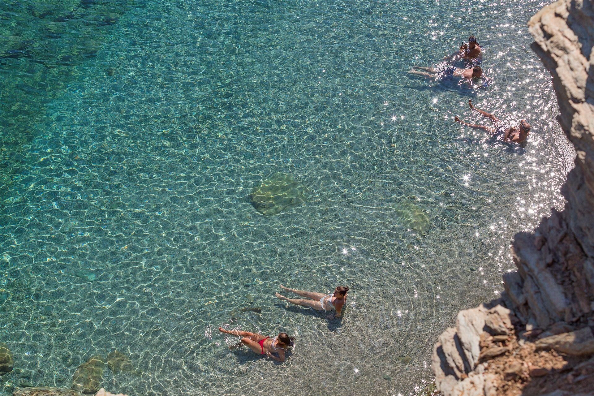 Ein klares, flaches Gewässer mit Schwimmern und Sonnenbadegästen an einem felsigen Ufer.