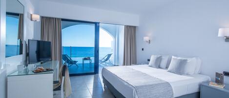 A modern hotel room with a large bed, a desk with a TV, and a balcony with a view of the sea.