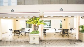A covered outdoor seating area with tables and chairs, a central palm tree planter, and a view of the building in the background. - iStay Hotel Monterrey Histórico (Monterrey)