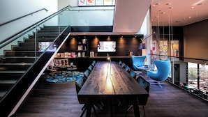 A modern interior with a wooden dining table, blue chairs, a staircase, and a bookshelf. - Motel One Wien-Westbahnhof (Vienna)