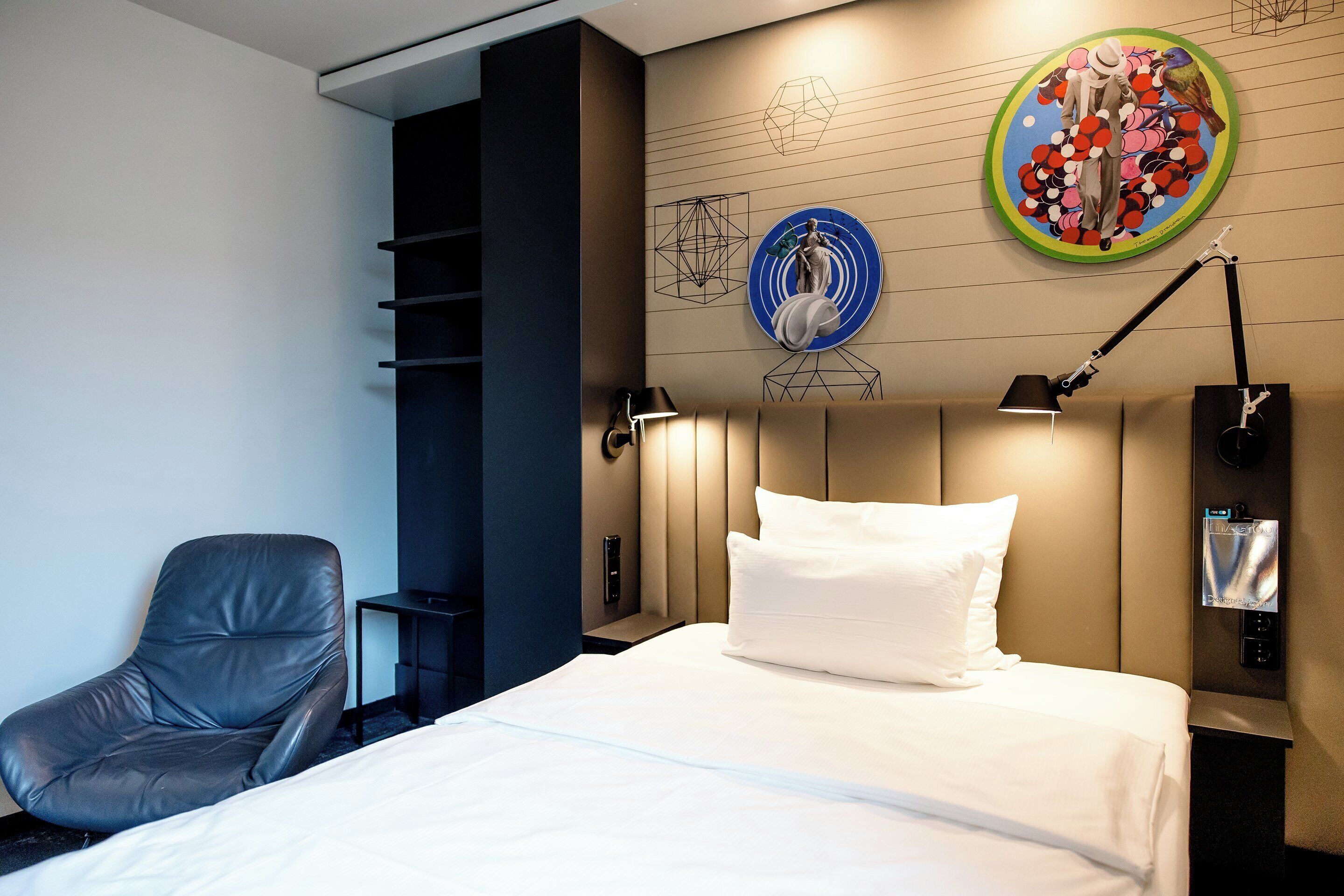 A hotel room with a bed, a chair, a wall-mounted lamp, and artwork.
