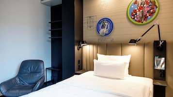 A hotel room with a bed, a chair, a wall-mounted lamp, and artwork.