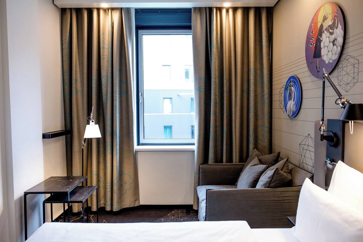 Una habitación de hotel con una cama, un escritorio, una silla, una lámpara de pie y una ventana con cortinas.